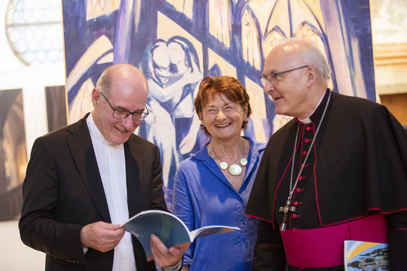 Dompropst Dr. Franz Frühmorgen, Dr. Maria Baumann und Bischof Dr. Rudolf Voderholzer stehen bei einer Ausstellungseröffnung vor einem großformatigen, blau‑weiß geprägten Gemälde. Dr. Franz Frühmorgen blättert in einem Katalog, während die beiden anderen nebeneinander stehen; Bischof Rudolf Voderholzer trägt geistliche Kleidung.